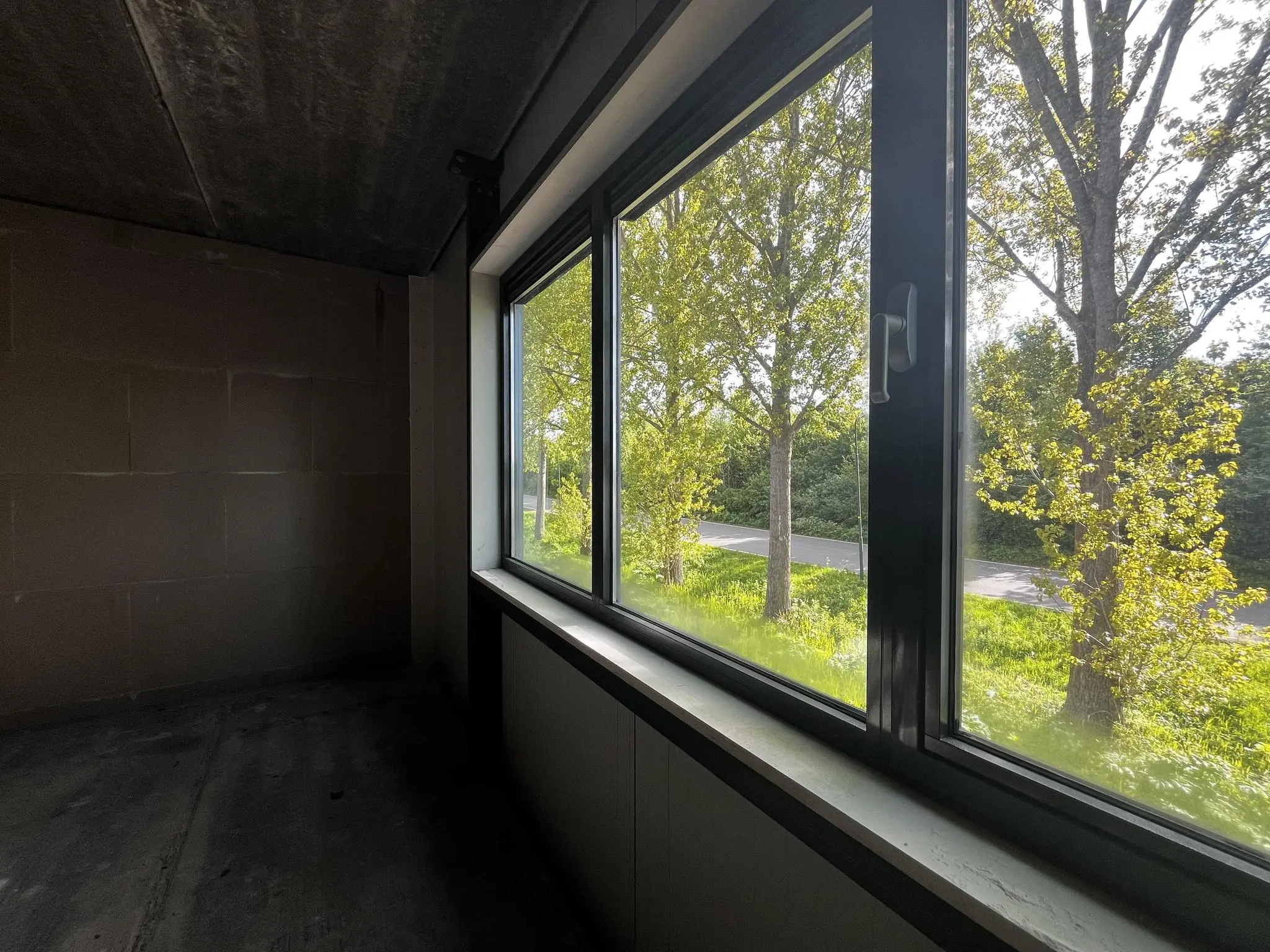 Uitzicht vanuit een lege kamer op Broekdijk-West met bomen en een weg onder een zonnige hemel.